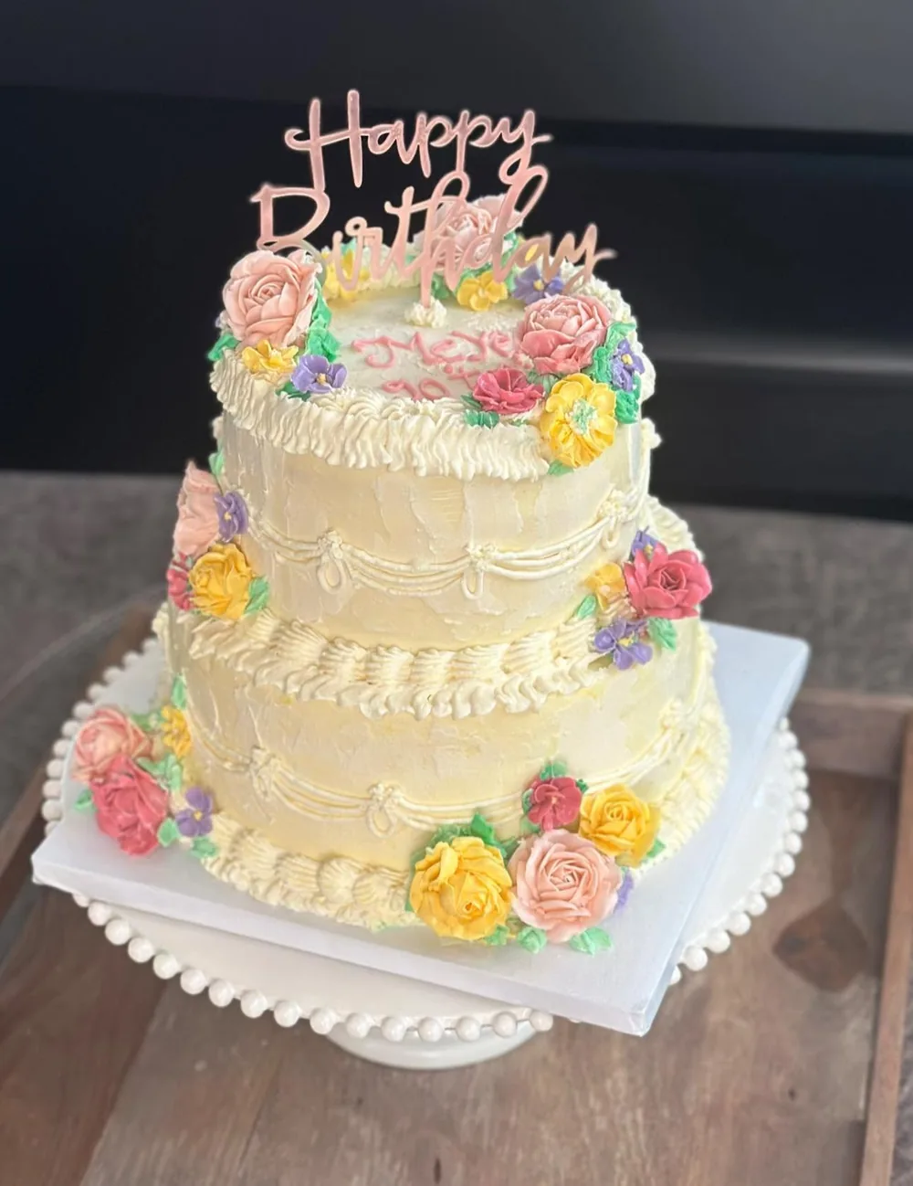 A two-tier cream cake with vintage piping, pink, yellow and lilac buttercream roses and a rose gold Happy Birthday topper